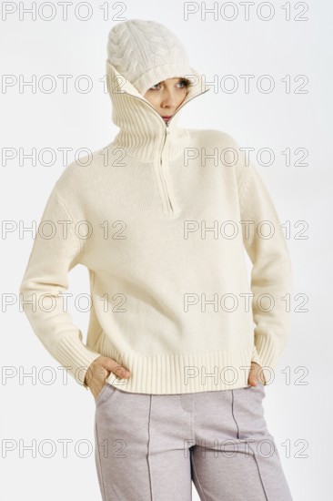 A woman stands in a neutral background wearing a warm knitted sweater and a hat. She looks relaxed with her hands in her pockets. The outfit is suitable for cold weather and casual settings
