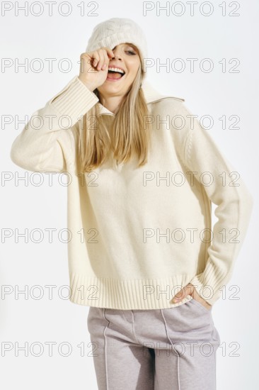 Playful woman pulls his hat over her eye. She is wearing a sweater and a warm pants. She smiles widely while playfully touching her face, showing a cheerful moment during a winter day