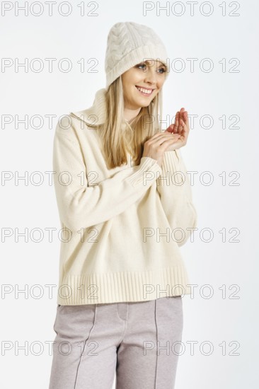 A woman stands with a smile, wearing a sweater and a hat. She clasps her hands together in front of her. The background is white, suggesting indoor lighting. The setting feels cozy and warm