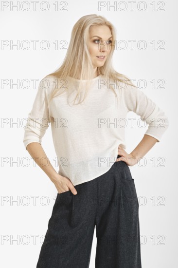 A woman stands confidently with one hand on her waist. They wear a light top and dark, loose-fitting pants in a minimal studio. The background is plain and focuses attention on their outfit