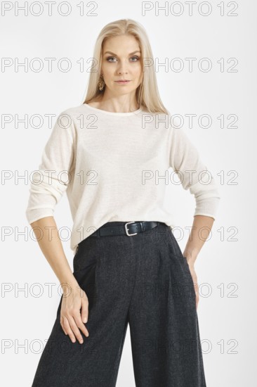A woman stands with a neutral expression in a studio. She wears a light top and gray wide pants. The background is plain and clear, focusing attention on her outfit and pose