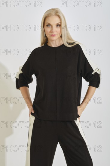 A woman stands with a confident pose wearing a comfortable black tracksuit with white stripes on both sides. The setting features bright lighting, highlighting her relaxed yet fashionable style