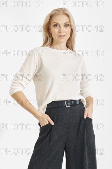 A woman poses with hands in pockets wearing a light shirt and dark pants. She has long blonde hair and stands in a well-lit studio with a simple backdrop. The scene focuses on her confident stance