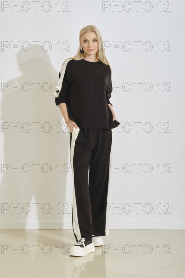 A woman stands confidently in a stylish black tracksuit featuring white stripes along the sides. She is in a well-lit indoor area with a simple background, projecting a modern fashion vibe
