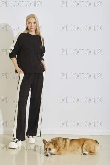 A woman stands in a sporty black tracksuit with white stripes while her dog lies comfortably on the floor beside her. The background is bright and simple, emphasizing their relaxed vibe