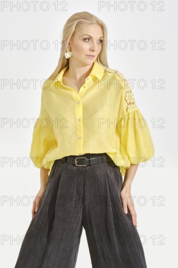 A woman poses confidently in a bright yellow blouse with intricate details, paired with relaxed black wide-leg pants. The elegant indoor setting showcases her fashion sense and vibrant personality