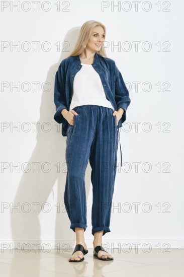 A woman is standing against a white wall in a bright room. She wears a blue pants and jacket with a white top. Her hair is long and straight. The floor has a shiny finish