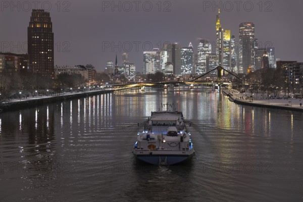 A ship sails towards the snow-covered and glowing Frankfurt banking skyline in the evening, Osthafen, Frankfurt am Main, Hesse, Germany