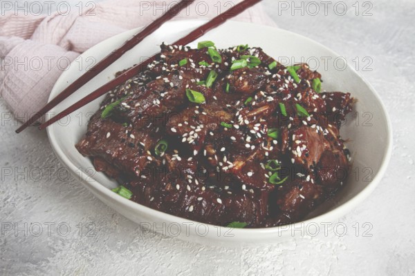 Crispy beef, with green onions and sesame seeds, in a bowl with chopsticks, on a textured surface, on a light background, no people