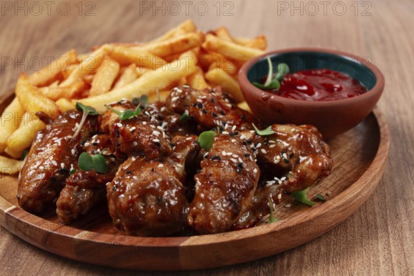 Barbecue chicken wings, sprinkled with sesame seeds, with French fries, and sauce, on a wooden plate, rustic style