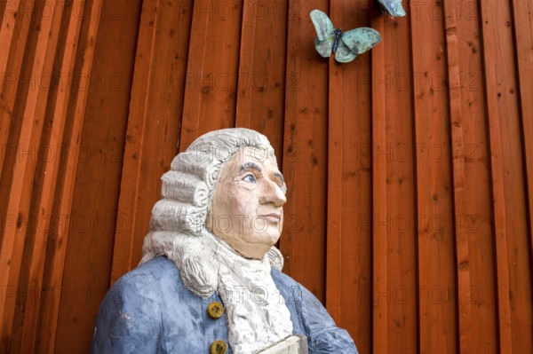 Wooden statue of botanist and founder of the binary nomenclature Carl von Linné with white hair and blue jacket in front of a wooden wall with a butterfly sculpture, Linnés Råshult, Älmhult, Kronobergs län, Småland, Sweden