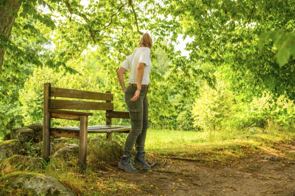Young woman in summer clothes standing next to a wooden bench in sunny forest, Linnés Råshult, Älmhult, Kronobergs län, Småland, Sweden