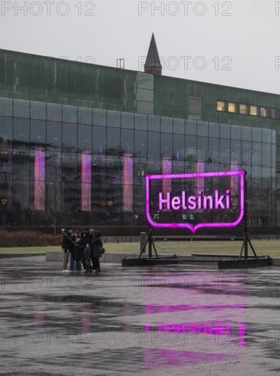 Tourists with selfie sticks take pictures of themselves in front of the glowing pink Helsinki city logo, square in front of Oodi Central Library and Cultural Center, Helsinki, Finland
