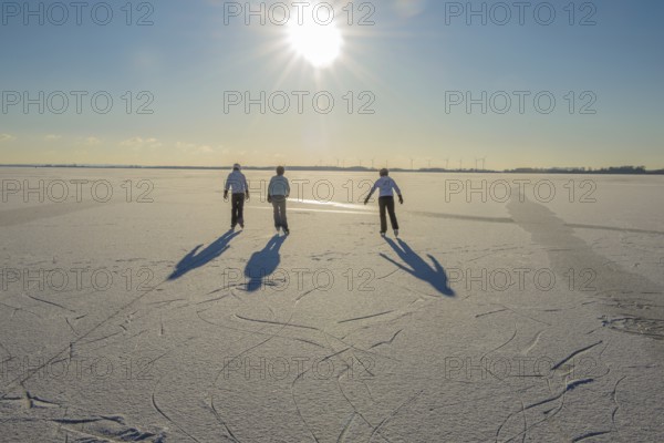 Three young woman skate on frozen Dümmer Lake under low sun, Lembruch, Lower Saxony, Germany