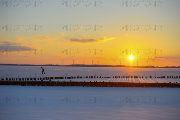 Ice skaters in front of sunset over icy landscape with wind turbines and intense colors on the frozen Dümmer Lake, Lembruch, Diepholz district, Lower Saxony, Germany