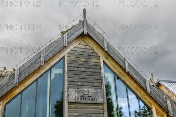 Modern building with wooden paneling and large glass windows under a cloudy sky, labelled as Café Doppingen on Hornborga Lake Hornborgassjön, Broddetorp, Västra Götalands län, Sweden