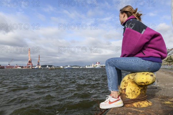 Person sitting on the shore looking at water and harbor cranes in an urban setting at Götaälv, Gothenburg, Västra Götalands län, Sweden