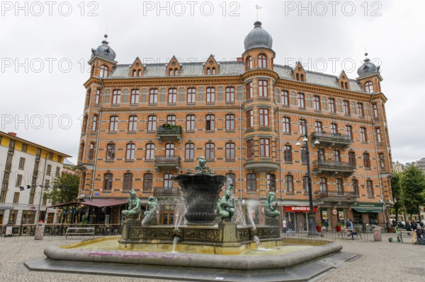 Historic brick building on Järntorget Square with the Järntorgsbrunnen also known under the Kümstler title The five continents with decorative elements and a fountain in the foreground on a cloudy day, the five naked bronze sculptures represent the 5 continents, Gothenburg, Västra Götalands län, Sweden
