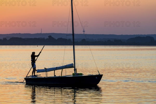 Sailing boat with person on calm Dümmer See brought into the harbor with a long stake at sunset, silhouette against orange sky, Lembruch, Dümmer See, Lower Saxony, Germany