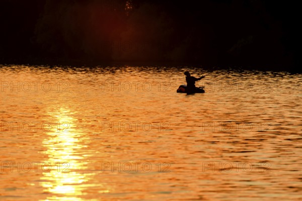Angler sport fisherman in his belly boat in orange-red lake at sunset, silhouette in calm water, relaxed atmosphere, Lembruch, Dümmer See, Lower Saxony, Germany