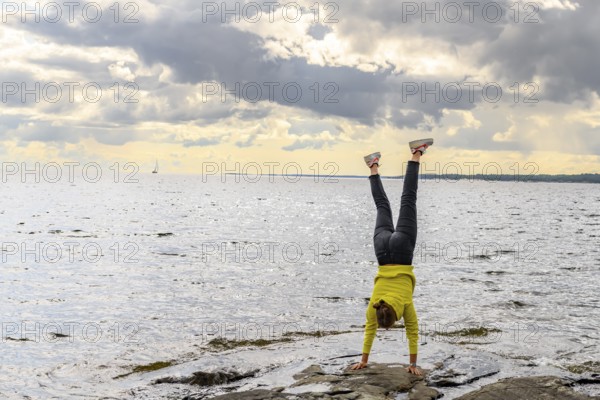 Young woman girl doing a handstand on rocky archipelago at Lake Vänern under cloudy sky, Värmlands län, Sweden