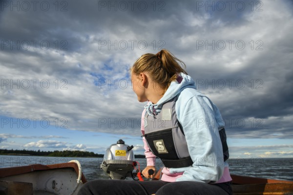 Young woman in a boat with outboard on the quiet lake Vänern, looking into the distance, the sky is cloudy, Vänern, Värmland, Sweden