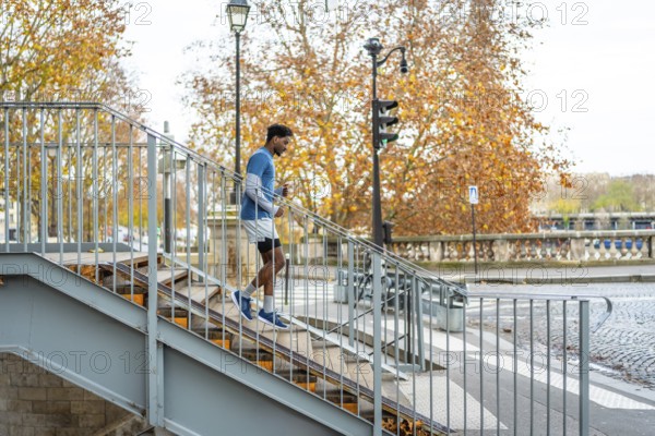 Energetic black man maintaining fitness by running down concrete stairs in an urban setting with autumn trees and a bridge in paris, demonstrating an active, healthy city lifestyle