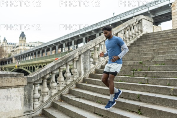 Young black man performing urban jogging and fitness training, actively descending large stone stairs in paris with a bridge over the seine river in the background