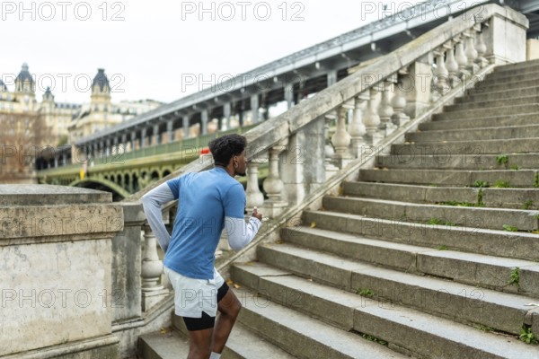 Young black man engaging in a fitness workout, actively running up historical stone stairs in paris with a bridge and buildings visible in the urban background