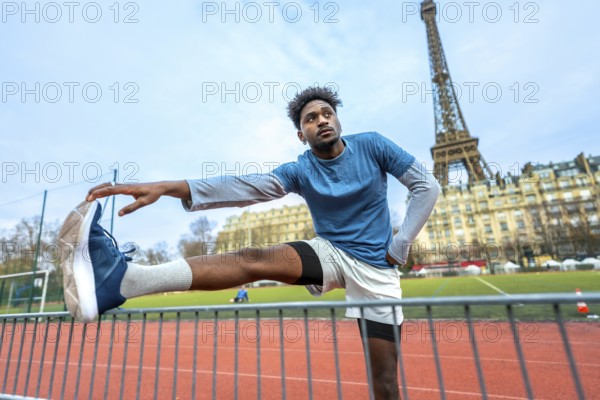 Young black man stretching legs on a paris running track before a workout, eiffel tower and haussmann buildings in the background, focused on urban fitness and dedication