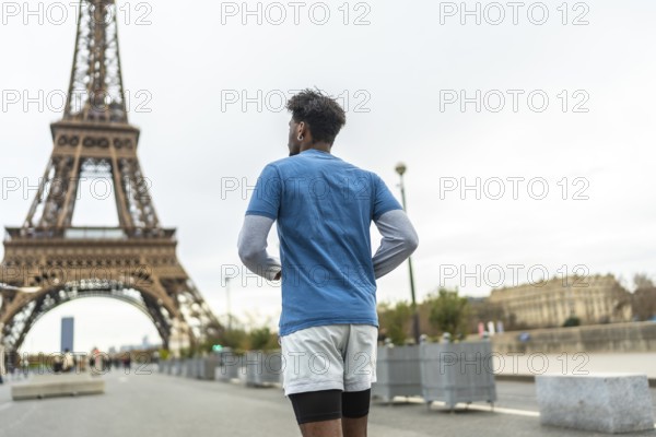 Adult man with dark skin running and training in the city of paris with the famous eiffel tower in the background on an overcast day, enjoying outdoor sport