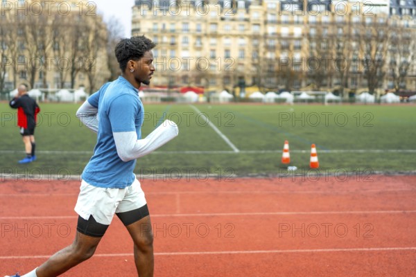 Sporty young black man running on an outdoor track in a stadium, focusing on fitness and urban training with buildings visible in the background, representing a healthy lifestyle