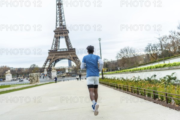 Young man in athletic wear jogging through a park pathway with the iconic eiffel tower prominently featured in the background on a cloudy day, focusing on fitness and tourism