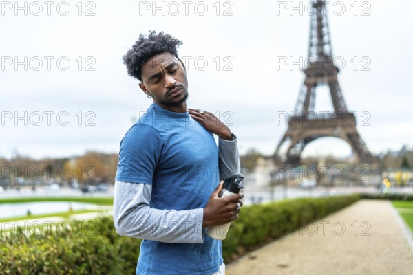 Young black athlete feeling neck pain and fatigue after exercising in a park, holding a water bottle with the eiffel tower visible in the background, embodying dedication to urban fitness