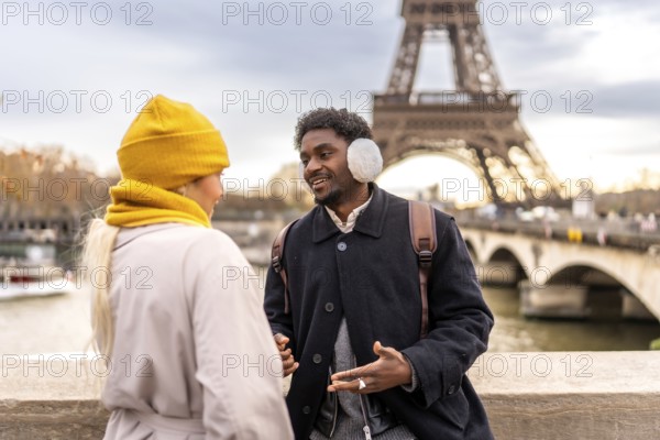 Diverse couple enjoying a romantic winter getaway in paris on a bridge by the eiffel tower, chatting and smiling as they sightsee along the seine during a city break