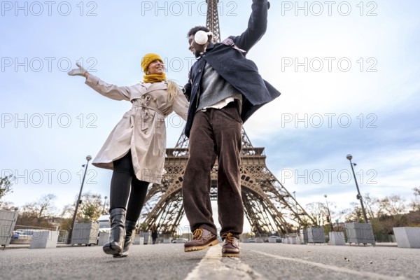 Happy diverse couple of travelers is enjoying a romantic vacation in paris, dancing with arms outstretched in front of the iconic eiffel tower, expressing joy and freedom