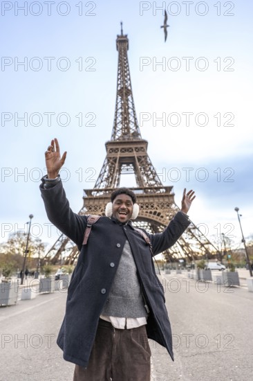 Young man with winter earmuffs raising arms in excitement and smiling widely while visiting the eiffel tower in paris, celebrating travel and tourism on a fun trip