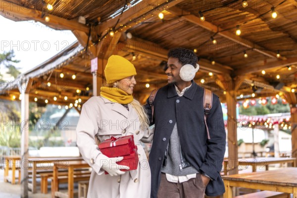 Diverse couple walking together and exchanging smiles in a festive outdoor christmas market in paris, carrying gifts and celebrating the holiday season