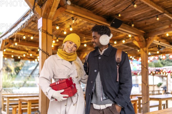 Young diverse couple walking together at a christmas market or winter fair, holding red gift boxes and smiling, celebrating holidays and special occasions