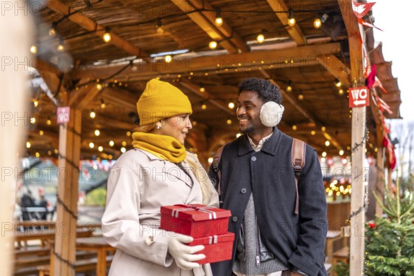 Young couple smiling at each other, walking and holding presents in red boxes at a festive outdoor christmas market in paris, enjoying a winter holiday together