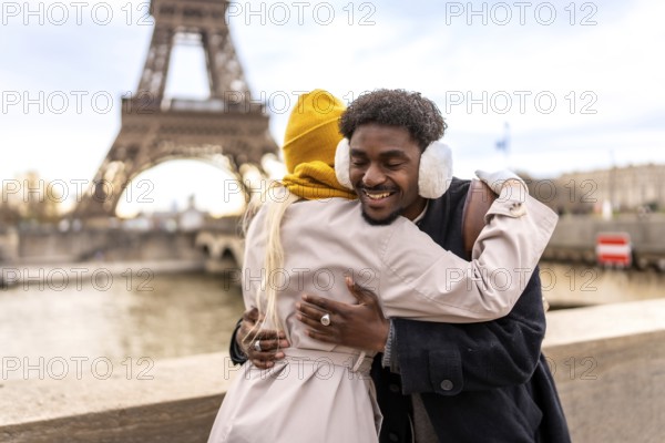 Happy young diverse couple hugging and smiling on a cold paris day, sharing warmth and romance with the eiffel tower softly blurred in the background