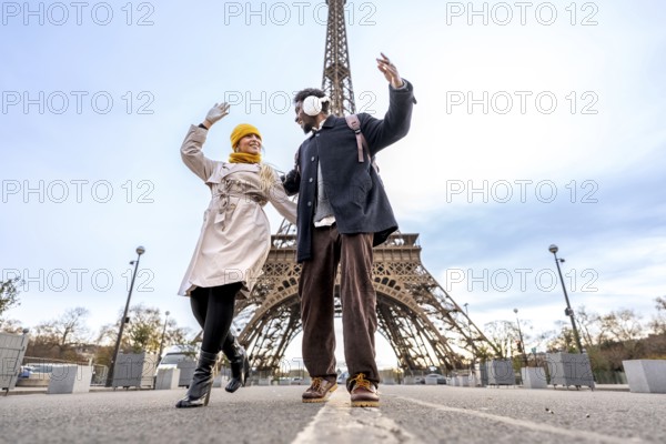 Happy young couple dancing with raised hands on a paris street by the eiffel tower, enjoying a romantic winter vacation together, smiling and full of joy and freedom