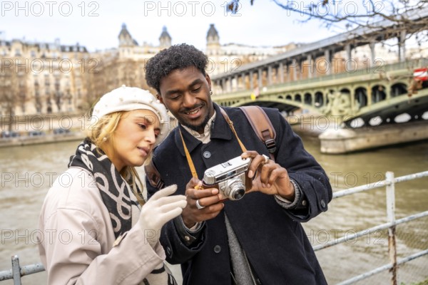 Young multiracial travelers enjoying a romantic city trip to paris, standing by the seine river and sharing a moment while looking at a vintage camera