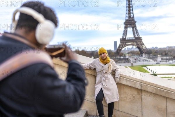 Couple traveling in paris. France. With a man taking a smartphone picture of his blonde girlfriend posing on a stone wall overlooking the iconic eiffel tower on a cloudy day. Creating travel memories