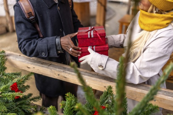 Man handing wrapped red christmas gifts to partner at an outdoor winter market, festive holiday exchange between a couple amid seasonal decorations and joyful crowd celebration