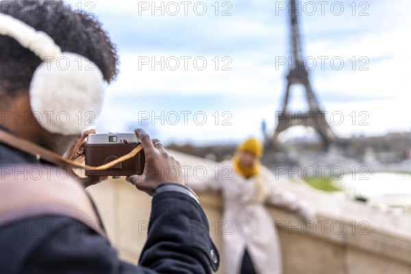 Young man wearing earmuffs and using a vintage camera to photograph his female partner posing with the iconic eiffel tower in the background, enjoying a romantic trip to a european city