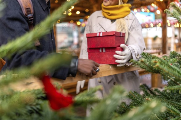 Diverse couple exchanging stack of red gift boxes, celebrating the holiday season at an outdoor christmas market, surrounded by festive decorations and evergreen branches