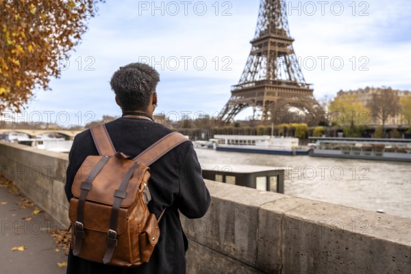 Person with travel backpack standing by the seine, gazing at the eiffel tower amid autumn foliage on a solo city trip in paris, enjoying urban riverside views and light breeze