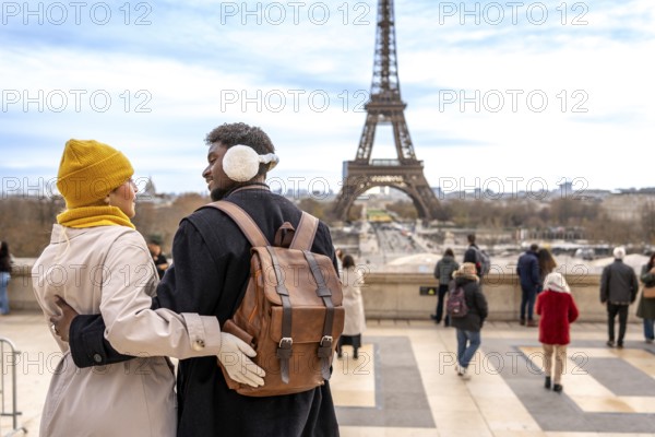 Young diverse couple embracing, traveling to paris, france, enjoying a romantic trip, sightseeing at trocadero with the eiffel tower in the background on a cold day