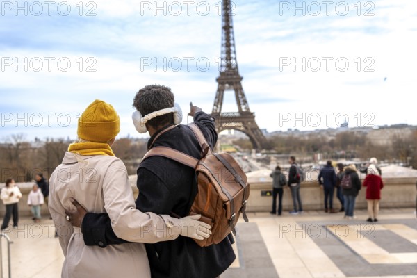 Young couple embracing and looking at the eiffel tower, the man pointing towards the landmark while enjoying a romantic winter travel experience in paris